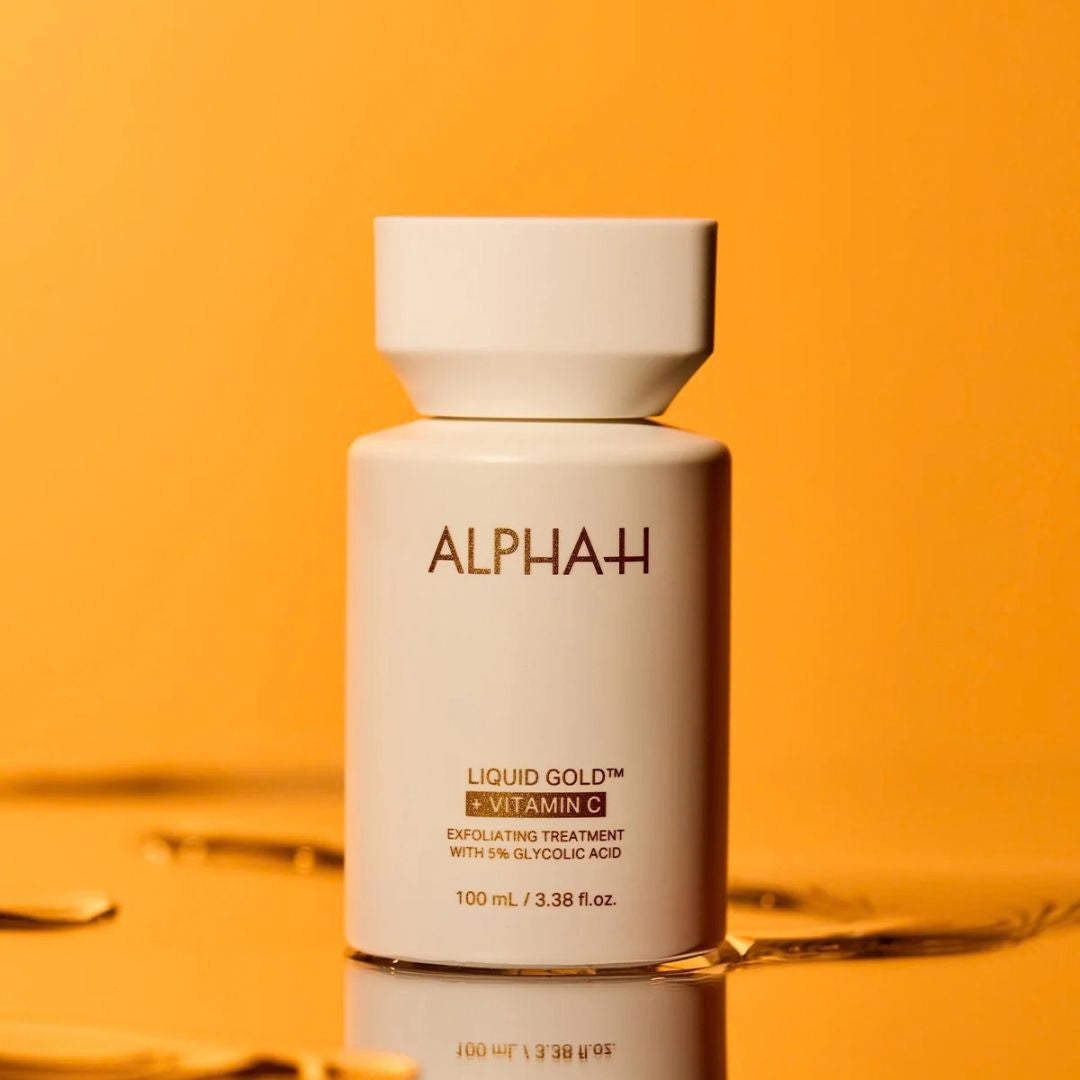 A white cylindrical bottle of ALPHA-H Liquid Gold + Vitamin C expolting treatment, set against a warm orange-yellow background with a reflective surface below.