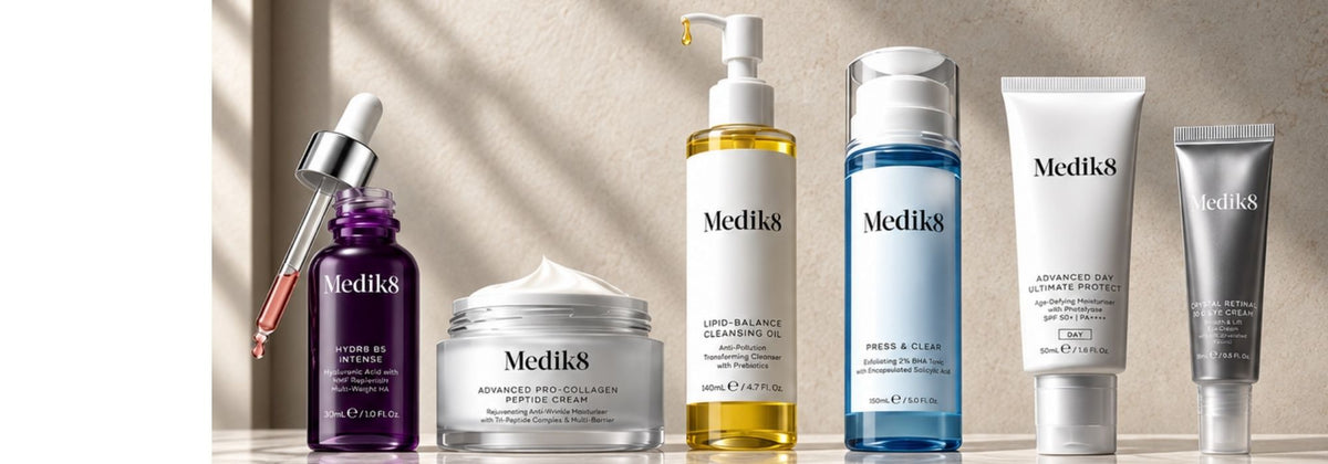 A lineup of Medik8 skincare products on a light beige background with soft diagonal shadows. The products include a purple dropper bottle, a white jar with cream, a yellow pump bottle, a blue bottle, a white tube, and a silver tube, all with the Medik8 brand name visible.