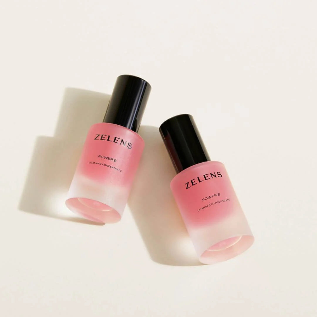 Two frosted glass cosmetic bottles with pink liquid and black caps, labeled ZELENS POWER B, arranged diagonally on a clean off-white background.