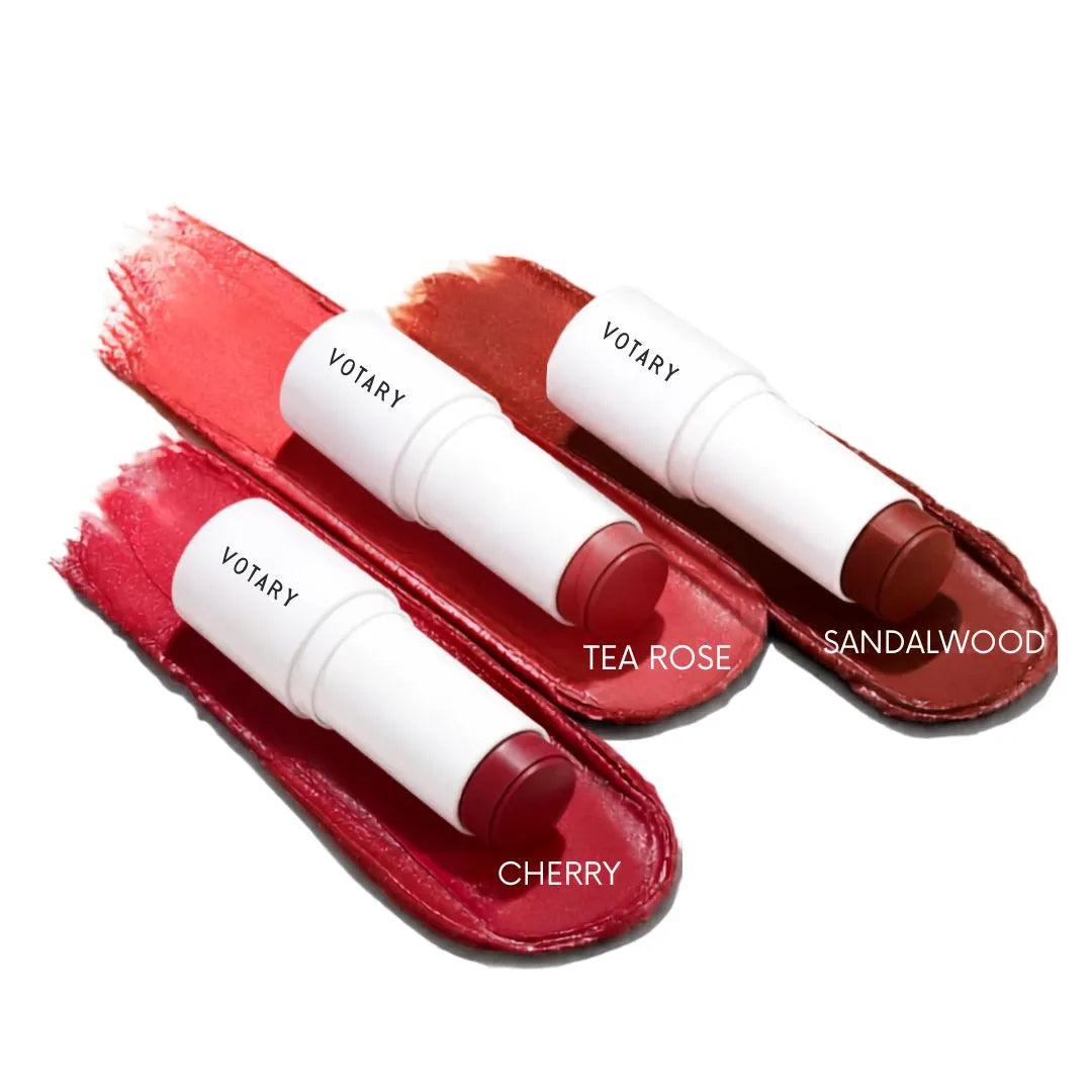 Two VOTARY lip balm sticks in white tubes with red caps, displayed on red textured swatches. One balm is labeled TEA ROSE and the other SANDALWOOD, with additional color labels CHERRY and RED.