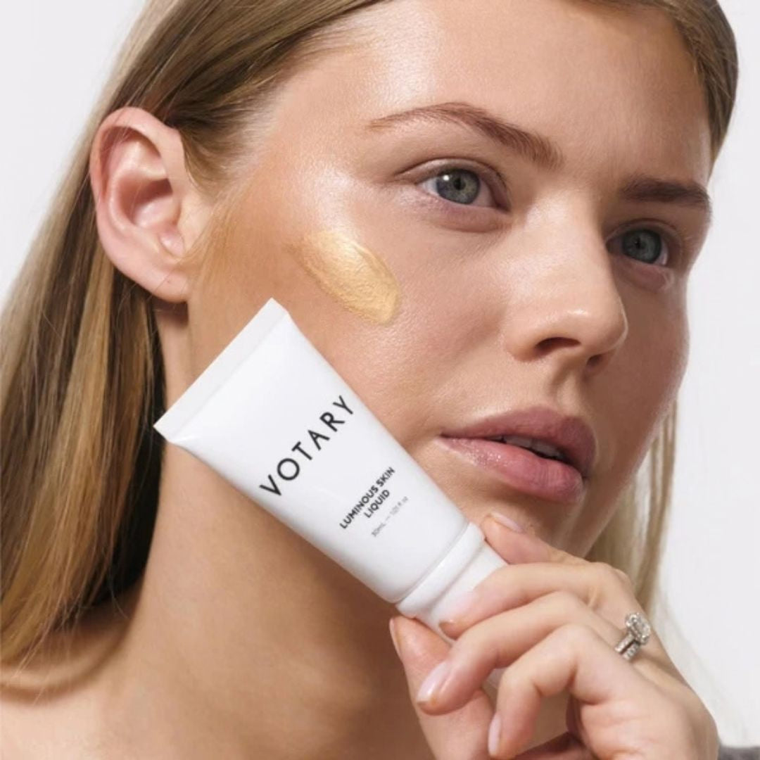 A close-up of a woman holding a white tube of VOTARY Luminary Skin BB Cream, with a dollop of the golden cream applied to her cheek against a clean white background.
