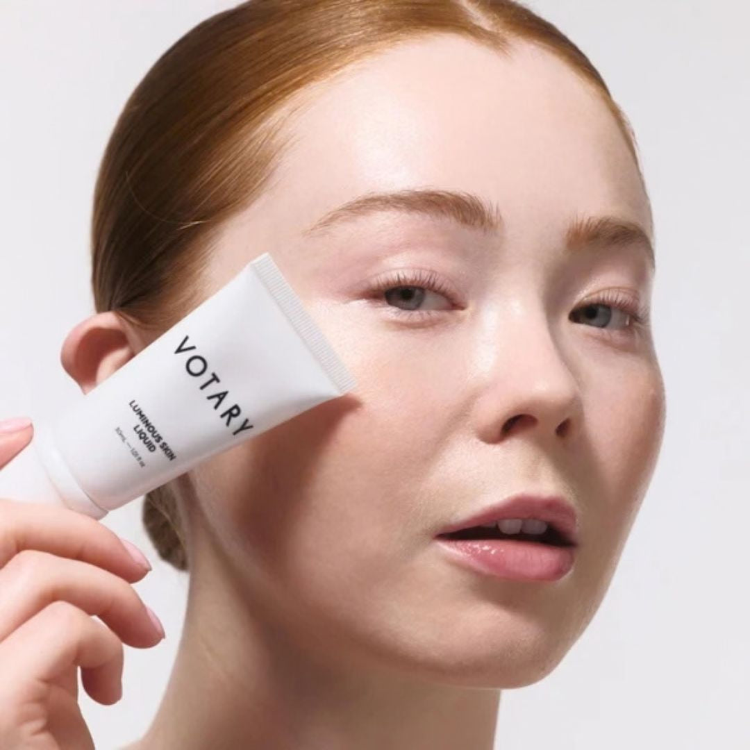 A close-up of a woman holding a white tube of VOTARY Lumière Rose skincare cream near her cheek against a clean light gray background.