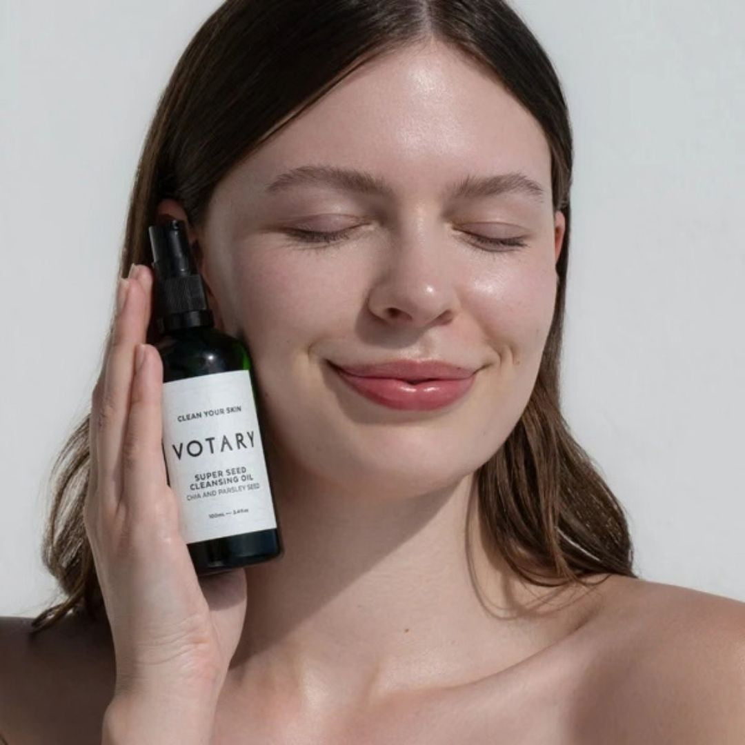 Votary Super Seed Cleansing Oil with chia and parsley seed held beside model with clean, radiant-looking skin