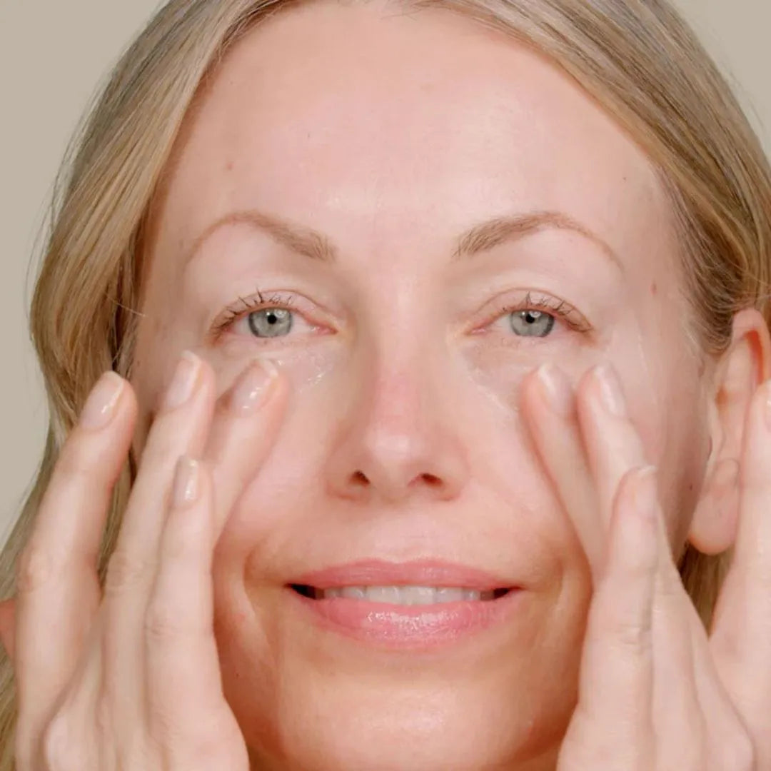 Close-up of a woman with blonde hair applying a clear eye cream to her under-eye area with her fingers against a soft beige background.