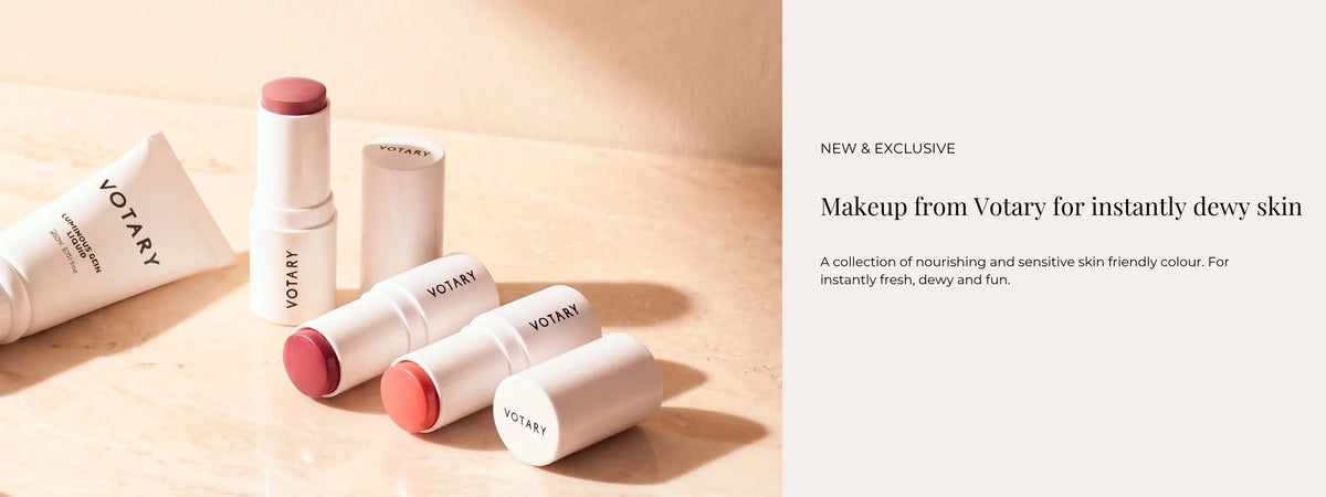 Votary makeup products arranged on a light beige surface beside a text panel that reads 'NEW & EXCLUSIVE Makeup from Votary for instantly dewy skin.' Visible products include white tubes and stick containers with pink and peach-toned caps or exposed makeup.