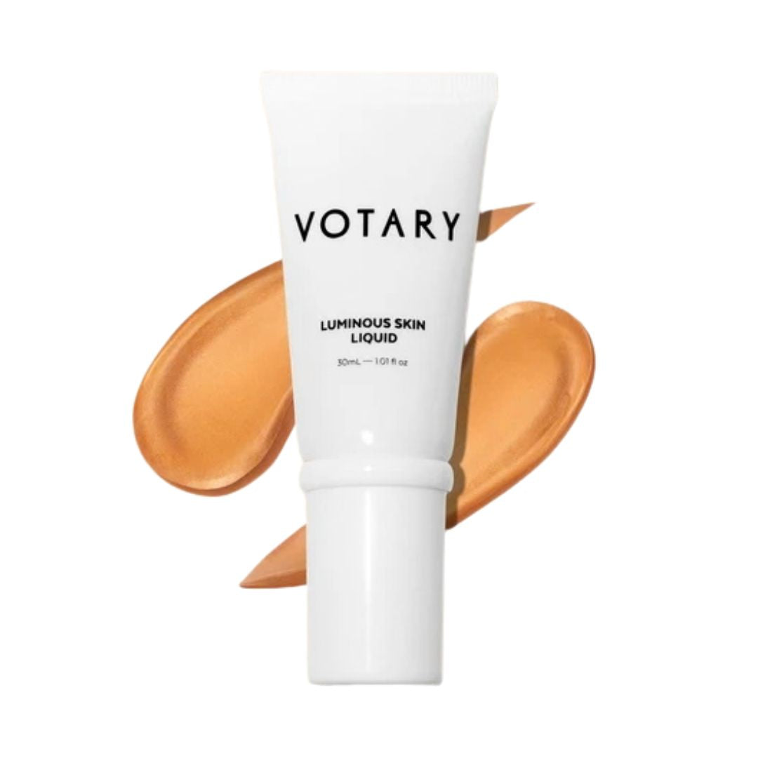 VOTARY Luminous Skin Liquid in a white squeeze tube, shown with two swatches of the warm beige liquid foundation behind it on a white background.