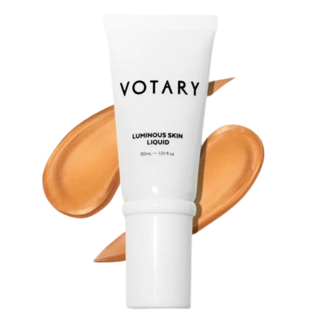 VOTARY Luminous Skin Liquid in a white squeeze tube, shown with two swatches of the warm beige liquid foundation behind it on a white background.