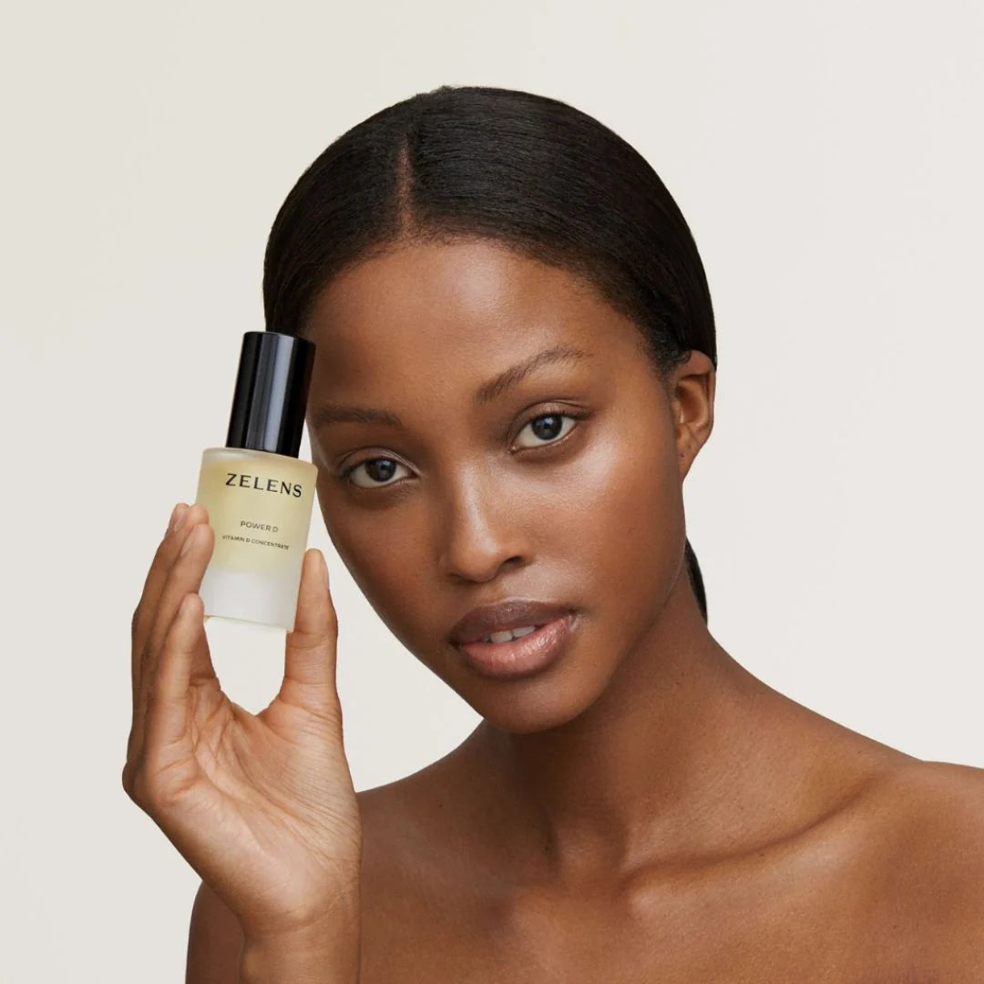 A woman holds a bottle of Zelens Probiotic Serum next to her face against a clean, light beige background.