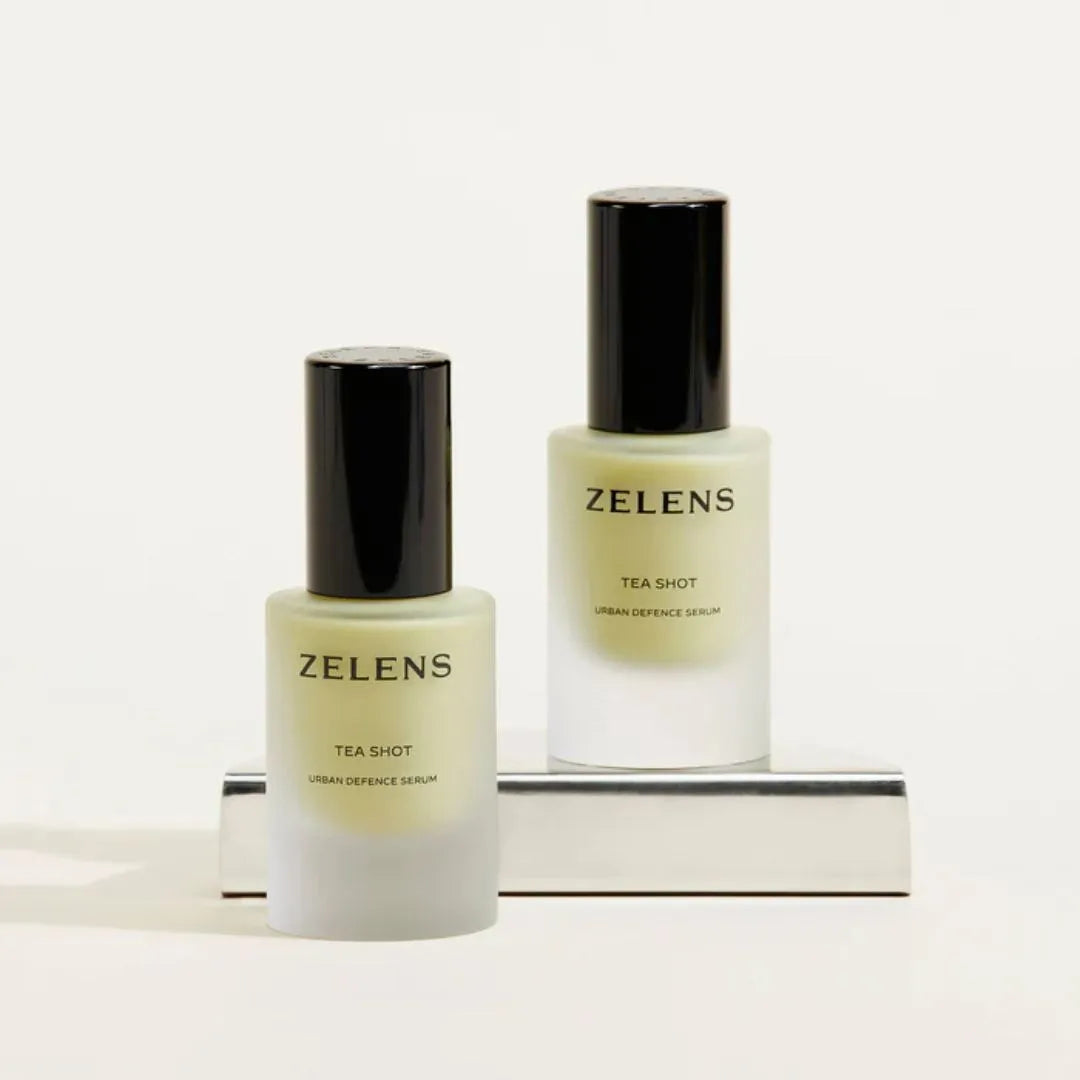 Two ZELENS Tea Shot Urban Essence Serum bottles with frosted glass and black caps, displayed on a minimalist white background with a reflective silver stand.