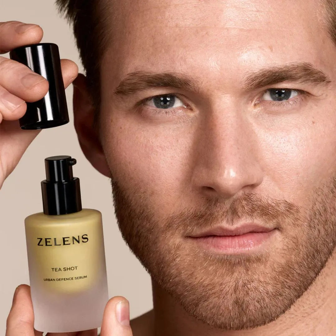 A close-up lifestyle shot of a man holding a bottle of Zelens Tea Shot Urban Defence Serum, with his hand lifting the black cap near his face against a warm beige background.