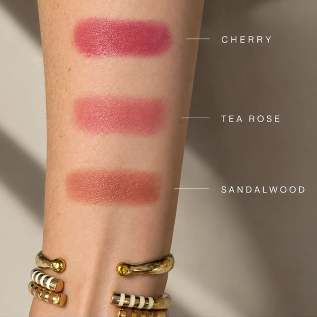 Three makeup swatches on a person's forearm labeled CHERRY, TEA ROSE, and SANDALWOOD, shown with gold and white striped bracelets against a soft neutral background.