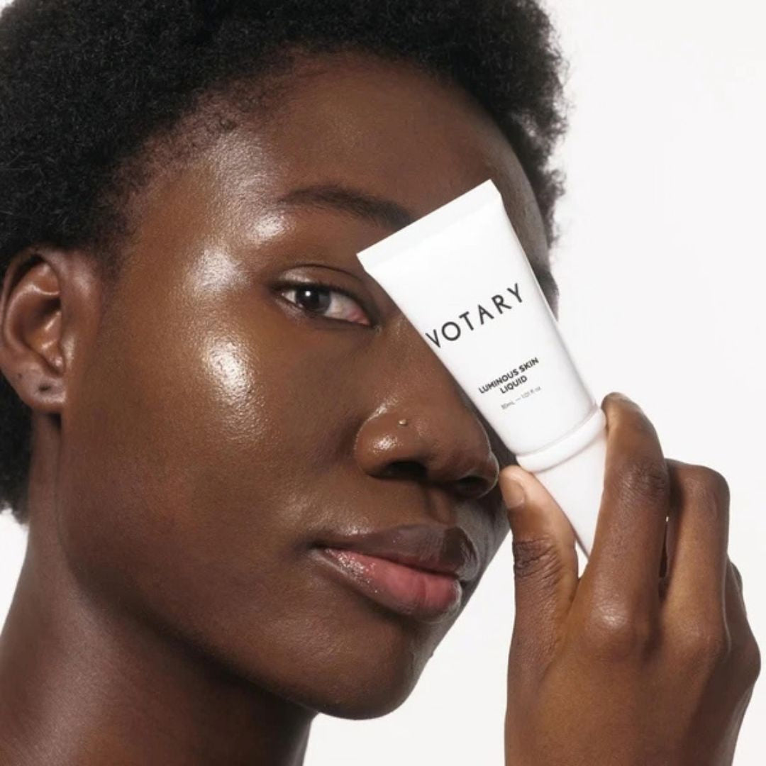 A close-up of a person holding a white tube of VOTARY Unmodified Skin Cream against their face, showing glossy, hydrated skin on a clean white background.