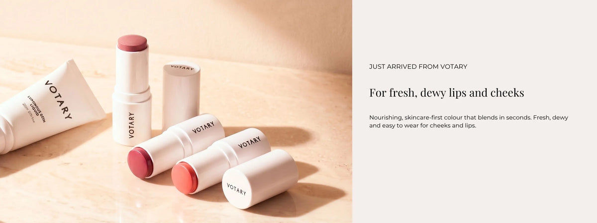 Votary lip and cheek balms in white tubes and sticks with pink and peach-colored balm tips, arranged on a light beige surface next to a text panel that reads 'JUST ARRIVED FROM VOTARY'.
