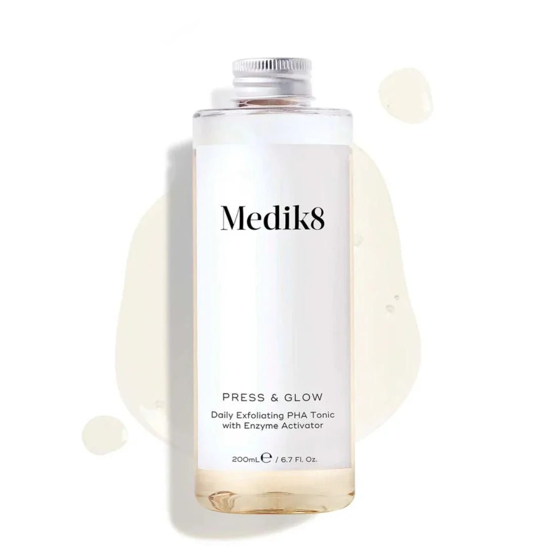 A clear plastic bottle of Medik8 Press & Glow Tonic with a white label and silver cap, set against a white background with soft beige liquid droplets.